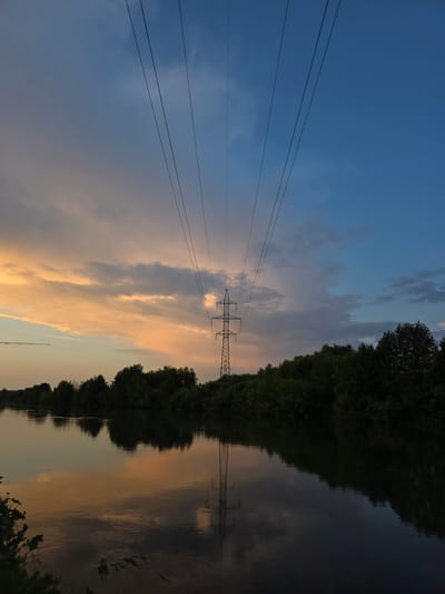 The image depicts a serene sunset over a river in Togashevo, Russia. The sky transitions from a soft orange and pink hue on the left, illuminated by the setting sun, to a deep blue on the right. Wispy clouds are scattered across the sky. A tall, metal electricity pylon stands prominently in the middle ground, with several power lines extending diagonally upwards towards the top of the frame. The pylon's reflection is visible in the dark, calm water of the river, which occupies the lower half of the image. Along the far bank of the river, a dense line of dark green trees forms a natural horizon. The overall atmosphere is peaceful and tranquil, characteristic of a rural landscape at dusk. There are no people or animals visible in the scene.