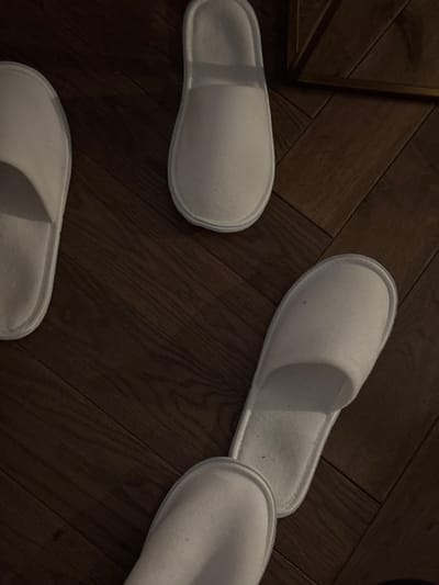 Three white hotel slippers are scattered across a dark, wooden floor with a herringbone pattern. A corner of a gold-framed object is visible on the upper left. The slippers are clean and appear to be unused, suggesting a hotel or spa environment. Light levels are subdued, emphasizing the contrast between the white slippers and the dark floor.