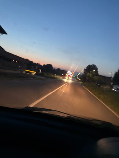 An outdoor, eye-level, slightly tilted shot captures a road at dusk, with a truck decorated with numerous lights driving away from the camera. The truck, positioned in the middle of the road, is illuminated by the colorful lights decorating its trailer and cab. The sky is a gradient of deep blue and orange hues, indicating twilight. The road, paved and with a white dividing line, curves gently to the right. To the left of the road, a building with lights visible in its windows is partially obscured by darkness and a roofline. Trees line the right side of the road, and streetlights are visible along the route. The foreground shows the dark interior of a vehicle, likely the one from which the photo was taken, with a windshield wiper visible. The overall scene is devoid of people. The location is Mezzana Sant'Antonio, Italy.