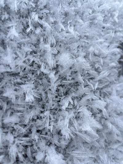 The image presents a detailed, close-up view of a dense formation of crystalline ice, consistent with hoarfrost or rime ice, observed in Карабоўскі сельскі Савет, Belarus. The frame is filled entirely with numerous individual ice crystals, primarily exhibiting needle-like, columnar, and feathery structures. These crystals are translucent to opaque white, with subtle grey and light blue tones, and vary in size and orientation, creating a highly textured, irregular surface. Darker, undefined areas are visible between the clusters of ice crystals, suggesting an underlying surface or depth within the formation. No human subjects, animals, or man-made objects are present. The scene depicts a static natural phenomenon associated with freezing temperatures.