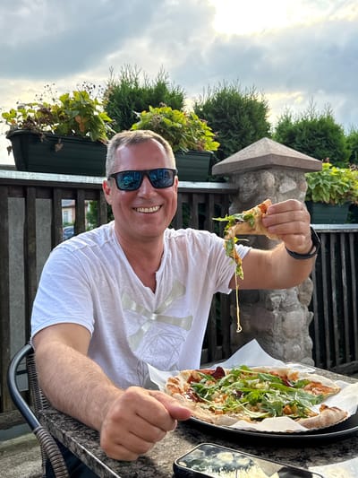 A middle-aged Caucasian man with short gray hair and sunglasses is sitting at an outdoor table, holding a slice of pizza. He is wearing a white t-shirt. In front of him on the table is a whole pizza. The background shows a wooden fence, some potted plants, and a stone pillar. The sky is cloudy. The image appears to have been taken in the city of Rūdiškės, Lithuania.