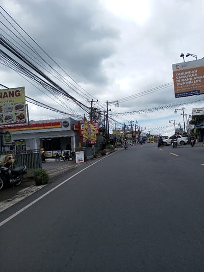 The image depicts an urban street scene in the city of Taohom, Laos, under a heavily overcast sky. A two-lane asphalt road forms the central element, flanked on the left by a paved sidewalk with a textured surface. On this sidewalk, a black motorcycle is parked facing left, and a small potted plant is present. Immediately adjacent to the sidewalk, behind a gray metal fence, stands a commercial building identifiable as a retail store, featuring a yellow canopy. A prominent yellow billboard displaying "NANANG NUSANTARA" with various food images is positioned in front of this building. Several individuals, including one wearing a hijab, are visible near the store's entrance, alongside multiple parked motorcycles. Red and yellow banner flags are affixed to poles near the storefront.
The road exhibits moderate vehicular traffic, with several motorcycles, some occupied by riders, and a white SUV visible in the mid-distance. One motorcyclist in the distance wears a light-colored helmet. Numerous utility poles supporting a dense network of overhead wires line both sides of the street and extend into the distance. On the right side, a large billboard displays Indonesian text: "TABUNGAN SADAYANA DI BANK AMAN DIJAMIN LPS." Additional buildings and streetlights are discernible along the street, receding into the background.