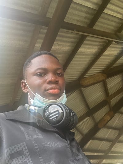 A single individual is photographed from the chest up, positioned indoors or under a shelter in Ozuaha, Nigeria. The subject, a male with dark skin, short dark hair, and a visible blemish on the right side of his nose, gazes directly at the camera. He wears a dark, short-sleeved collared shirt with a subtle geometric pattern. A light blue surgical mask is lowered, resting below his chin, and black "Wireless One" over-ear headphones are around his neck. Above and behind the subject, the ceiling consists of a corrugated metal roof supported by dark wooden beams.