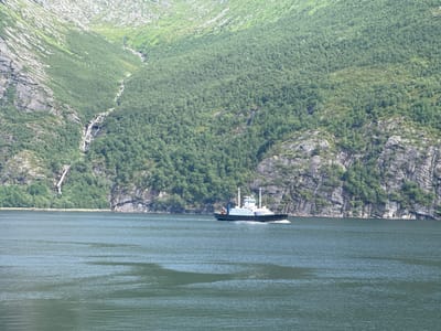 A ferry boat travels on a calm, dark green body of water. The ferry is white with a dark hull and has several decks and a white superstructure. It is moving from left to right. In the background, a steep, lush green mountainside rises dramatically from the water. A narrow waterfall cascades down the rocky, sparsely vegetated upper portion of the mountain. The lower slopes of the mountain are densely covered in green trees. The sky is not visible in the frame. The scene depicts a natural landscape with a man-made vessel. The location is identified as Inndyr, Norway.