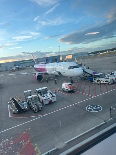 The image provides an aerial view of an active airport tarmac, likely taken from a terminal window, with a large passenger aircraft as the central subject. The time of day appears to be late afternoon or early morning, characterized by a bright, partly cloudy blue sky with hints of warmer light on some clouds.
The main subject is a Wizz Air Airbus A320 family aircraft, identifiable by its distinctive white fuselage with a prominent purple/pink tail and belly, and the "WIZZ AIR" logo clearly visible on its side. The aircraft is parked at gate "20," as indicated by the large "20" painted within a circle on the concrete tarmac.
Several ground crew members, wearing high-visibility vests, are visible around the aircraft, engaged in servicing activities. Various pieces of ground support equipment surround the plane: a mobile set of stairs is connected to the forward passenger door, a pushback tug is positioned at the nose gear, and multiple other service vehicles, including baggage loaders and smaller service trucks (one with a red and white checkered pattern), are stationed near the wings and fuselage. These vehicles and personnel suggest the aircraft is undergoing turnaround procedures, such as boarding, deboarding, or preparation for departure.
The tarmac features clear white and red painted lines marking parking areas and safety zones. In the background, other airport structures, such as hangars and possibly other parked aircraft, can be seen, extending towards the horizon under the broad sky. The overall scene depicts a busy operational environment at an airport, consistent with the specified location of Luton, United Kingdom.