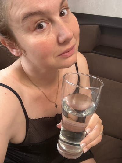 A woman is holding a glass of water, with her head tilted to the side and her eyes wide open, looking at the camera. She is wearing a black top with lace detailing and a delicate gold chain with a cross pendant. The background appears to be a domestic setting, possibly a living room, with a brown couch visible. The lighting suggests it could be daytime. The woman has light skin and her lips are slightly parted. Her nails are neatly manicured in a pale pink shade, and she is wearing a ring on her finger. The image is framed tightly on her face and upper body, with the glass of water prominently featured in the foreground. The overall mood of the image is somewhat inquisitive or surprised, due to the woman's direct gaze and wide eyes. There is no visible text in the image.