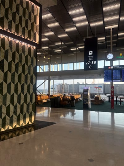 This image captures an interior view of a gate or lounge area within an airport terminal at Charles de Gaulle Airport (CDG) in Roissy-en-France, France.
In the left foreground, a prominent column is clad in a geometric pattern of dark green and light-colored marble, forming an illusion of three-dimensional cubes. The vertical edges of this column are illuminated by integrated strip lighting. The floor in the immediate foreground is composed of polished light-colored marble or tile, exhibiting reflections of the overhead lighting.
The central and background portions of the image feature a spacious waiting area bordered by a wall of large windows. Through these windows, an airport tarmac is visible, including the fuselage and wing of a parked airplane connected to a jet bridge. An advertisement for "TotalEnergies" is discernible on a structure outside.
Within the lounge, rows of brown or tan cushioned benches and individual seating units are arranged on a dark green carpeted floor. Multiple individuals are present, with one person clearly visible seated on a bench, looking downwards, next to a black rolling suitcase. Other passengers are indistinctly seated in the background.
Suspended from the ceiling, which features a grid of metallic panels and integrated strip lighting, is a black sign with white text reading "Portes Gates 登机口 27-38" and a left-pointing arrow, accompanied by an airplane icon. To the right of this sign, an analog clock with "ROLEX" branding is mounted, and adjacent to it, a digital departures board displays flight information. An ATM machine, labeled "ATM," stands in the midground. A decorative chandelier with exposed bulbs hangs in the mid-air section. The environment is brightly lit by a combination of natural daylight entering through the windows and overhead artificial lighting.