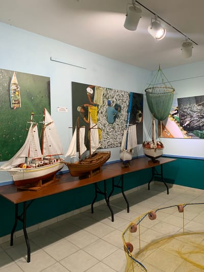 The image depicts an indoor exhibition space, likely a museum or cultural center, in Boca del Río, Venezuela. The walls are painted a light blue, and a dark teal-colored wainscoting runs along the lower portion of the walls. Track lighting illuminates the space from the ceiling.

The primary subjects of the exhibition are model boats displayed on a long, wooden table. These models range in size and detail, with some featuring white sails and intricate rigging, while others are simpler wooden designs. One particularly prominent model has the inscription "AMSHI 2016" on its hull. Behind the table, large framed photographs are mounted on the wall, showcasing scenes related to fishing and maritime activities. One photograph shows an aerial view of a boat on green water, while others depict people working with fish and fishing nets.

Suspended from the ceiling by a green rope is a large, circular fishing net, also acting as an exhibit. In the foreground, on the tiled floor, a yellow fishing net is partially visible, secured by a frame and adorned with pink floats. The overall impression is an educational or historical display about local fishing traditions.