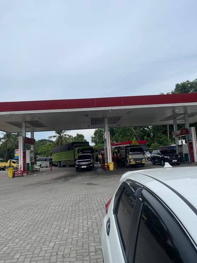 The image depicts a scene at a gas station in Glee Gogo, Indonesia. The perspective is from inside a white car, with its windshield and side window visible in the foreground. The gas station has a red and white canopy over several fueling stations. Multiple vehicles, including cars and trucks, are lined up, suggesting a queue for refueling. One prominent vehicle is a green truck. There are also people visible, presumably gas station attendants and customers. The ground is paved with interlocking bricks. Lush green trees and palm trees are visible in the background, indicating a tropical or subtropical environment. The sky is overcast, suggesting a cloudy day. The time of day appears to be daytime. Text is visible on some of the vehicles and gas station equipment, but it is largely illegible in the provided image. One truck has a license plate that appears to read "BK 3477 PI". The overall impression is a busy but ordinary day at a gas station in Indonesia.