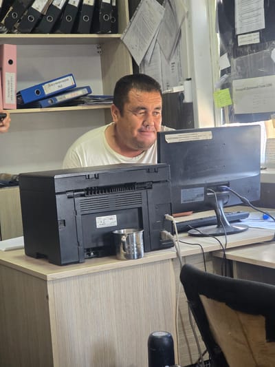The image shows a man in a white t-shirt looking at the camera with a neutral expression. He is standing in an office or storage area. Behind him, there is a desk with a computer monitor, a printer, and various papers. A black office chair is partially visible. On the left side of the image, there are shelves filled with binders. On the right, there is a light-colored wooden cabinet. The overall setting appears to be a workplace with an emphasis on document management. Some visible text includes "ДОКУМЕНТЫ," "ПРОЕКТ ТЕХНИЧЕСКИЙ ПРЕДЕЛ," "Накладной Дорожный Газли 1240," and brand names like "SAMSUNG" and "Canon." The lighting suggests it is daytime.