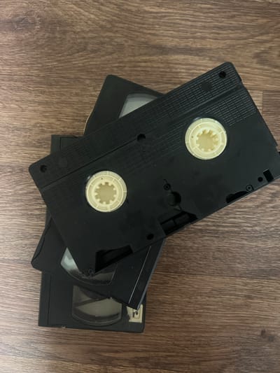 The image shows two stacked VHS tapes on a wooden surface. The top VHS tape is black and is positioned diagonally across the bottom tape. It features two cream-colored spools visible on its surface. The bottom VHS tape is also black and is placed horizontally. The wooden surface appears to be a floor or table with a visible grain pattern. The lighting suggests an indoor environment. There are no people or any indication of the location being in Amirovo, Russia.