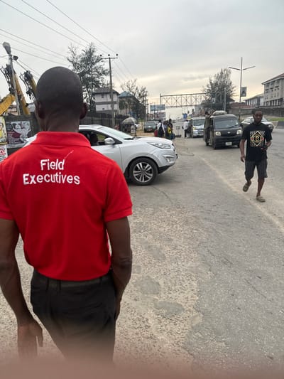 A street scene showcases a man in a red "Field Executives" polo shirt facing away from the camera, partially obscuring a white car. The street is busy with traffic including black cars, vans, and numerous pedestrians. A man in a black patterned shirt walks down the middle of the street. Overheard power lines and billboards frame the view. The sky is overcast and the scene appears to be set in an urban area, possibly a city in Africa.