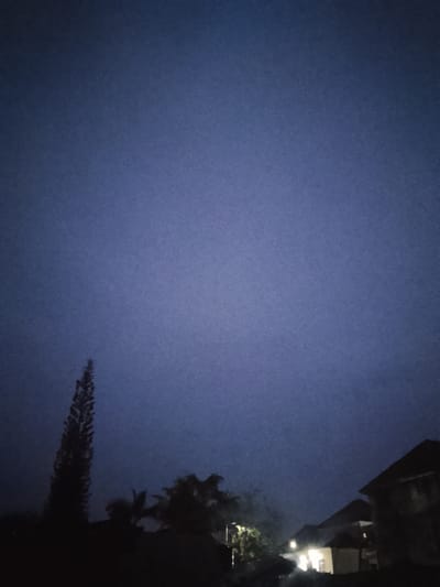The image depicts a dark, twilight scene in Rumuokwuta, Nigeria. A tall, dark silhouette of a tree or large plant dominates the left side of the frame. In the lower right corner, the outlines of buildings with illuminated windows suggest a residential area. The sky is a deep, uniform blue, indicating the transition from day to night or pre-dawn darkness. The overall atmosphere is calm and quiet, with no discernible human activity. The low light conditions obscure finer details, creating a sense of mystery and stillness.
