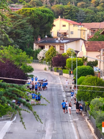 This high-angle, vertical media file captures a lively street scene in Passignano sul Trasimeno, Italy, likely during late afternoon or early evening due to the warm, golden light filtering through the trees and illuminating the buildings. The weather appears to be pleasant, with clear skies.

The scene is dominated by a residential street winding uphill, bordered by lush greenery and multi-story houses with terracotta roofs. Numerous people are visible, many dressed in blue shirts and shorts, suggesting a group event or a local sports team. A large group of individuals, predominantly young men, are walking up the street, some in pairs or small clusters. A few people are gathered on a stone bench by the side of the road, engaged in conversation.

Notable details include the vibrant trees, including large coniferous trees and dense deciduous foliage, some with deep purple leaves. The houses exhibit architectural styles common in the region, with balconies and awnings. A few street lamps are present, and a clothesline with laundry is visible on one of the houses. One of the blue shirts worn by a person walking up the street has white lettering, possibly a team name or logo, but it is not clearly legible. Another person is wearing a blue shirt with white lettering that appears to say "CAMIONCINO".

The overall atmosphere seems relaxed and social, with people casually walking and gathering. The presence of multiple groups of people, many in similar attire, suggests a community event, a parade, or a gathering related to a local festival or sports activity. The scene is set on a paved street with white lane markings. The sunlight creates soft shadows, indicating the sun is lower in the sky.