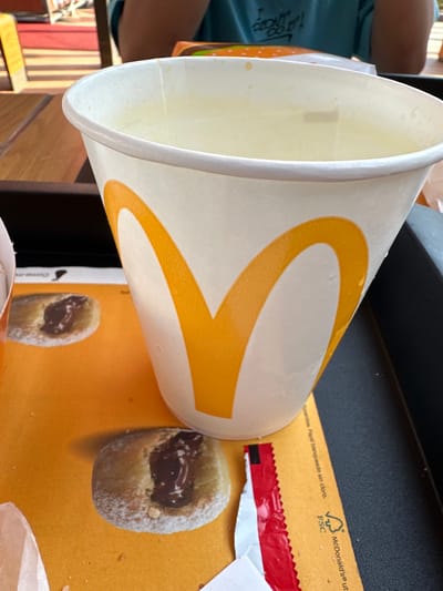 This image shows a close-up view of a white paper cup with the McDonald's logo, filled with a pale yellow liquid, likely a beverage. The cup is placed on a tray that features an orange surface with a printed design. On the orange surface, two small pastries are visible. One pastry, in the foreground, appears to be a doughnut dusted with powdered sugar and filled with a dark chocolate or jam filling. A red and white wrapper, possibly from a condiment or food item, is also present near the pastries. The background shows a person's arm and torso, wearing a teal t-shirt. The setting appears to be an indoor or outdoor seating area, possibly a McDonald's restaurant. The presence of the McDonald's branding strongly suggests the location is a McDonald's establishment. Given the context of McDonald's, it is plausible that this scene is located within the city of Sedaví, Spain, where a McDonald's restaurant exists. The lighting suggests it is daytime.
