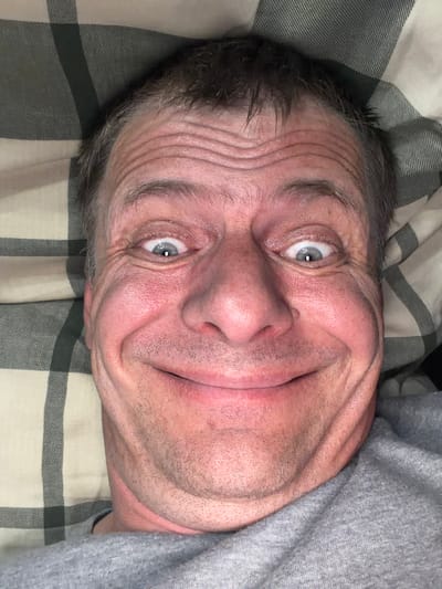 The image is a close-up, selfie-style portrait of a middle-aged white man with thinning brown hair. He is lying down, with his head resting on a pillow that has a plaid pattern of beige and dark gray. The man has a wide, exaggerated smile, with his eyes open wide and a slight squint. His forehead is creased with wrinkles, and his cheeks are flushed. He is wearing a gray t-shirt. The image does not contain any information about the city of New Britain, United States of America.