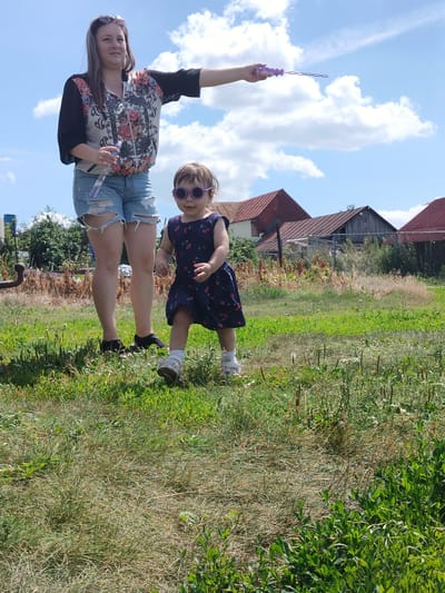 A woman is standing in a grassy field with a young child. The woman is wearing a black and floral print bomber jacket, ripped denim shorts, and black shoes. She is holding a bottle of water in her left hand and a bubble wand in her right hand, with bubbles in the air. The child is wearing a dark blue dress with cherries, sunglasses, and white shoes. They are in a rural setting, with wooden buildings in the background and trees and vegetation surrounding them. The sky is blue with white clouds. The overall scene suggests a casual outdoor activity on a sunny day. The location is identified as Блохино, Russia.