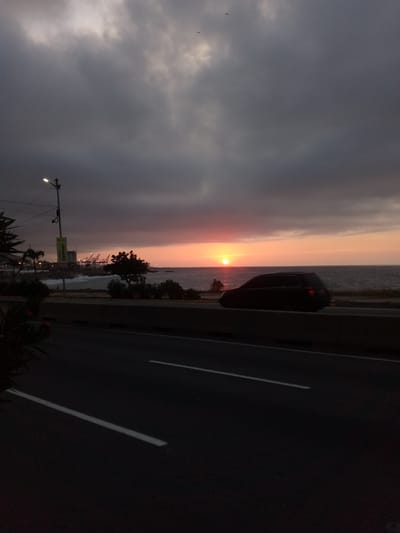 The image depicts a coastal scene at sunset in La Guaira, Venezuela. A dark-colored vehicle is in motion on a road parallel to the coastline. The sun is setting over the ocean, casting an orange glow on the horizon and through the cloudy sky. In the background, port infrastructure, including cranes, is visible along the coast. A lamppost stands near the road, and some trees and foliage are present. The road itself has white lane markings. The overall impression is one of a twilight coastal drive.