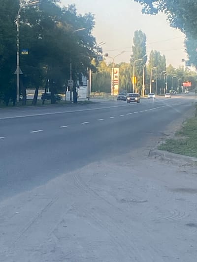 The image depicts a roadside scene in Pavlohrad, Ukraine. The perspective is from a moving vehicle, looking towards the right side of the road and slightly upwards. The primary subjects are a road with lane markings, several cars, trees lining the left side of the road, and a roadside embankment on the right.
The road appears to be a two-lane highway with a white dashed line separating the lanes. Several cars are visible on the road, with some appearing to be moving. The trees are dense and green, suggesting a temperate season, likely summer or late spring, given the bright sky. The sky itself is bright and appears clear, indicating it is daytime, likely late afternoon due to the angle of the light. The roadside on the right is covered with a rough, greyish surface, possibly gravel or dirt, with some vegetation growing near the edge.
Notable details include traffic lights and streetlights along the left side of the road. A billboard or large sign with red and white stripes is visible further down the road. There is also a smaller sign with what appears to be a Ukrainian flag. The overall scene conveys a sense of everyday travel or commuting in a semi-urban or suburban environment. No specific event or emotional tone is apparent; the image captures a typical roadside view. No discernible text is visible in the image.