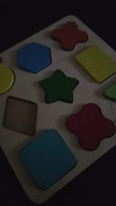 This is a close-up, high-angle shot of a wooden shape sorter puzzle. The puzzle is mostly empty, with several brightly colored geometric shapes placed in their corresponding slots. From top left, clockwise: a blue hexagon, a red clover-like shape, a yellow rectangle, a green star, a red clover-like shape, and a blue square. There is also a yellow circle and a brown square-ish shape visible in other slots. The background is dark and out of focus. The image does not provide any information about the city of Sannikovo, Russia.