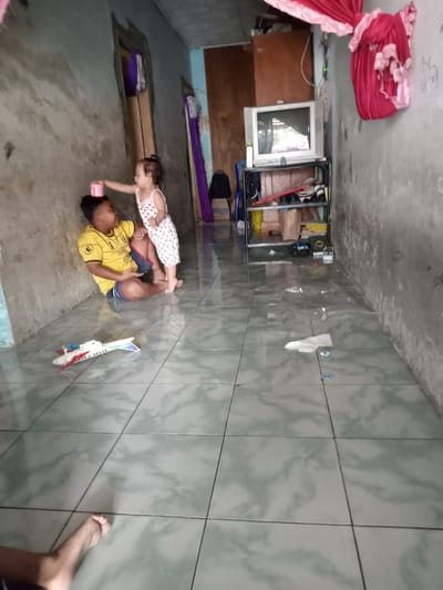 Location: Bungur, Indonesia.
Two children are interacting on a tiled floor. A younger female child in a white and red patterned dress is standing, placing a pink cup on the head of an older male child seated on the floor. The boy is wearing a yellow shirt. A toy airplane lies near the children. A television on a metal shelf stands against the back wall, next to a wooden cabinet. The walls are bare and appear aged. Red fabric hangs along the top of the right wall.