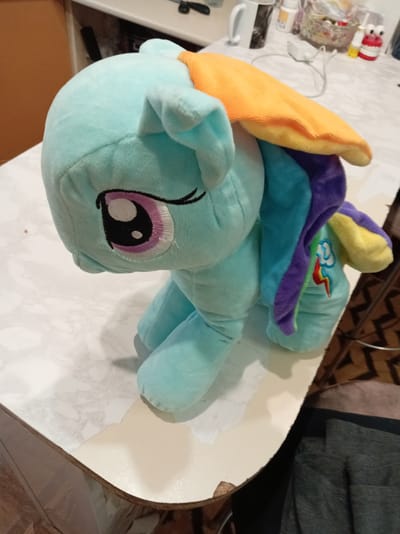 This is a close-up, medium shot of a light blue plush toy depicting Rainbow Dash, a character from My Little Pony. The toy is positioned on a table with a marble-like pattern. Rainbow Dash is facing left, with its head lowered. Its eye is large and purple with a black pupil. The toy has orange, blue, and purple hair, and a rainbow cloud symbol is visible on its flank. The background is blurred, showing a hint of a shelf with various objects, including a white mug and what appears to be medication bottles. The table surface shows signs of wear and tear, with peeling edges. The scene appears to be indoors, in what could be a domestic setting in a suburban or rural area of Belarus. There is no discernible activity taking place beyond the toy being placed on the table. The lighting suggests it could be daytime.
