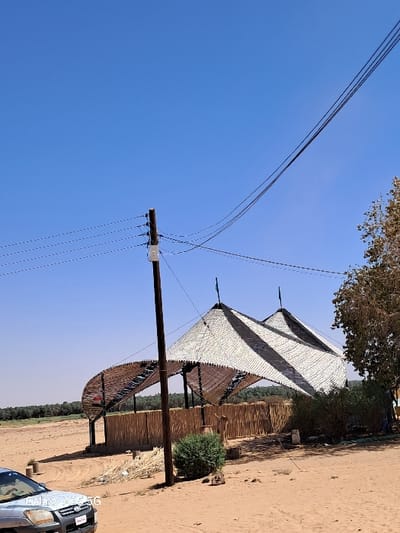 The scene is an open, sandy area in Karima, Sudan (18.54, 31.831) under a clear, bright blue sky. A large, white and black striped tent-like structure, supported by wooden poles, stands in the center of the image. The tent is constructed of woven material and has a distinct curved, arched shape. A wooden power pole with multiple power lines running overhead stands in front of the structure. There are several trees in the background, a few of which are bare and others with lush green foliage. In the foreground, a portion of a silver car is visible, parked in front of a small, dense bush. The sand is slightly uneven and appears dry.  The scene has a deserted feel.