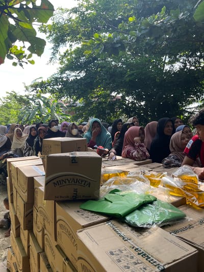 In Pulodolang, Indonesia, a group of people, mostly women wearing hijabs, are gathered outdoors, seemingly waiting in line. They are positioned behind stacks of cardboard boxes, many of which are labeled "MINYAKITA" and appear to contain cooking oil. Several pouches of cooking oil are visible on top of some of the boxes. The environment is partly shaded by trees, with the sky visible through the leaves. Some individuals are wearing face masks. The overall impression is of a distribution event, likely for food aid.