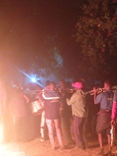 A group of people stand illuminated by a fire, several holding trumpets and one with a drum. The illumination casts a warm glow, contrasting with the surrounding darkness. One person wears a jacket with "DRILL SPECIALIST" on the back and patterned pants. Another wears a pink cap. Trees are visible in the background. The scene suggests a musical performance or gathering taking place outdoors at night.