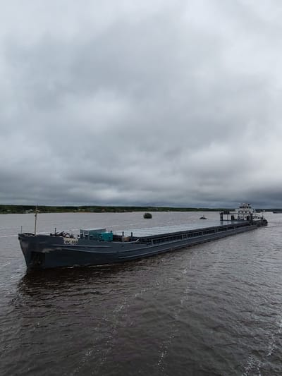 The image shows a large, gray barge moving through a body of water. The barge is long and rectangular with a superstructure at the rear and the Cyrillic letters "CФС-555" visible on the front. The water is choppy, indicating some wind. The sky is overcast with dense, gray clouds. A treeline is visible in the distance, and a small cluster of buildings is discernible among the trees on the left. This scene is in Shestovo, Russia.