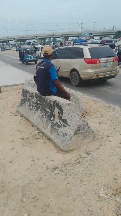 A man in a blue and black shirt and tan cap sits on a large, grey, concrete block in Effurun, Nigeria. He holds a cell phone in his hand. The block is on a sandy area next to a road. A tan Toyota Sienna minivan is partially visible on the road. A blue and black tricycle vehicle is driving away. An overpass bridge is in the background, and an advertising billboard is also visible.