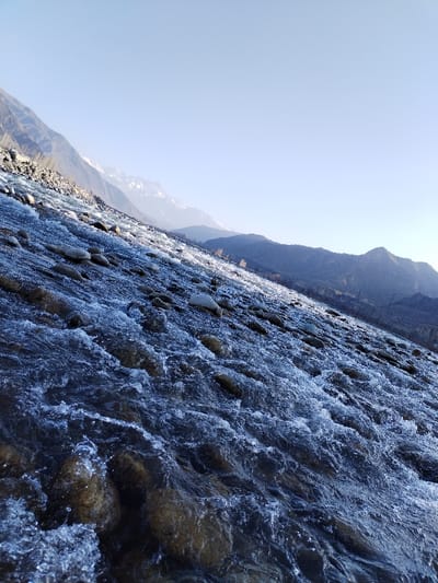 The image displays a mountainous natural landscape within Qoraqiya, Uzbekistan, centered on a rapidly flowing watercourse. Dark blue water, characterized by significant turbulence and white froth, moves over a bed of varying-sized, smooth rocks. Portions of the water's surface and the rocks are covered with white and light blue formations consistent with ice and slush. To the left in the background, a steep, rocky mountain face rises. A distant mountain range, partially obscured by haze and featuring visible snow-capped peaks, extends across the central background. Lower, rugged hills are present on the right background. The upper section of the image shows a clear, light blue sky. No human figures, animals, or man-made structures are visible. The scene is illuminated by natural light, indicating daytime conditions in a cold environment.