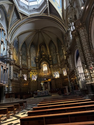 The image displays the grand, ornate interior of a basilica or cathedral, viewed from within the nave, looking towards the altar and upwards towards the vaulted ceilings.
The primary subjects are the elaborate architectural and decorative elements of the church. Dominating the upper and left portions are high, ribbed vaulted ceilings, richly adorned with intricate gold trim, painted sections, and detailed moldings. The walls below are covered with numerous frescoes or large paintings depicting religious scenes, framed by luxurious gold and plasterwork. In the middle distance, a magnificent, heavily gilded altar or reredos serves as a central focal point, brightly lit and featuring statues and sacred imagery.
On the right side of the nave, rows of dark wooden pews are visible, indicating seating for a congregation. In the lower left foreground, a substantial pipe organ with numerous polished silver pipes is prominently positioned. A large, arched window with multiple panes on the far left allows natural light to enter the vast space. The floor, partly visible, features a geometric pattern of dark and light tiles.
No specific activity or event is taking place; the church appears empty of people, suggesting a moment of quiet or a time outside of a religious service.
The time of day is likely daytime, with natural light from the window complemented by artificial lighting that highlights the intricate details and artworks, casting a warm, reverent glow. The lavish use of gold, detailed carvings, and extensive religious iconography points to a significant Catholic church. The scale, architectural style, and opulent decoration create an atmosphere of immense grandeur and spiritual solemnity. There is no visible text.