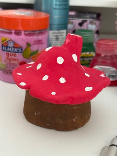 A close-up, slightly angled view shows a red-and-white mushroom figurine resting on a white surface. The mushroom's cap is bright red with scattered white spots. Its stem appears to be brown. The background contains a variety of crafting supplies, including a jar of pink Elmer's slime and other unidentified containers. The location is not indicated.