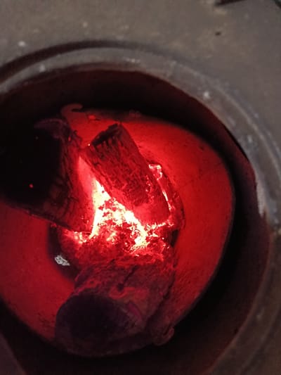 The image depicts a contained wood fire from an overhead perspective, situated in Vlado Trichkov, Bulgaria. Several pieces of wood are in an advanced state of combustion within a dark, circular metal or ceramic combustion chamber. A focal point of bright orange flames and glowing embers is concentrated towards the center-right, with incandescent wood radiating intense red and orange light. Ash is intermixed with the burning fuel. The interior walls of the chamber are illuminated by the fire's reddish-orange glow, contrasting with the dark, unlit outer rim of the opening visible at the top and left.