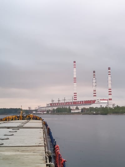 The image shows the Konakovo Power Station in Konakovo, Russia, from the perspective of a platform on the water. Three tall smokestacks with red and white stripes are prominent features of the power station. The main building of the power station bears the inscription "КОНАКОВСКАЯ ГРЭС" (Konakovo GRES). The sky is overcast, and the water is calm. In the foreground is a concrete platform with some yellow machinery and red ropes visible. A treeline is visible to the left side of the frame.