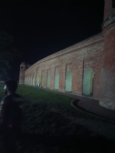 In the city of Bagerhat, Bangladesh, at night, a brick structure with arched openings stretches into the darkness. The building's facade, constructed from reddish-brown bricks, is illuminated by artificial light, revealing a series of evenly spaced arched alcoves along its length. To the left, a silhouette of a person is visible, facing away from the camera and towards the structure. The foreground consists of a grassy area, with a curved pathway visible in front of the building. The sky above is dark and featureless.