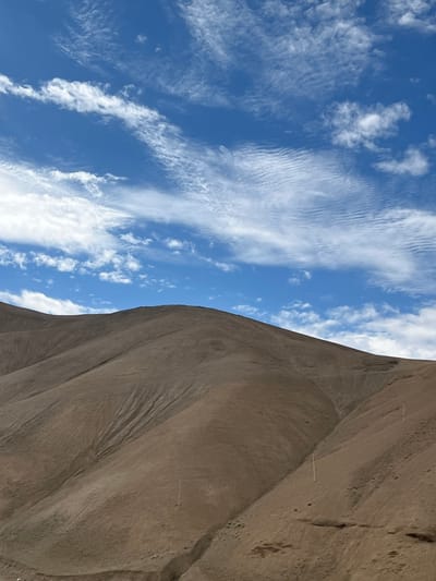 The image shows a vast, arid mountain landscape under a bright blue sky dotted with wispy white clouds. The hills are a uniform, light brown color, with subtle variations in tone suggesting gentle slopes and undulations. Deep ravines and gullies carve their way down the mountainsides, indicating erosion from water or wind. A few thin, white poles are visible, standing vertically on the slopes, possibly marking trails or infrastructure. The overall impression is one of a remote, natural environment, possibly in a high-altitude region. The location is specified as Wakha, India, a region known for its mountainous terrain. There are no people or animals visible in the image.