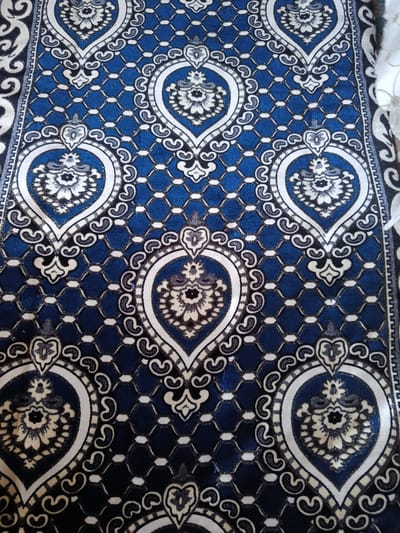 The image displays a close-up view of a blue rug with an intricate white and black floral pattern. The rug features repeating symmetrical designs, including paisley shapes and stylized flowers enclosed within teardrop-like borders. The overall texture appears to be velvety, with a subtle sheen that catches the light. The pattern is dense and detailed, creating a rich visual tapestry. The rug is laid out flat, with no visible people or surrounding environment other than a hint of a white surface to the right, suggesting it is indoors. The specific location within Гулбог 1, Tajikistan, cannot be determined from the image alone.