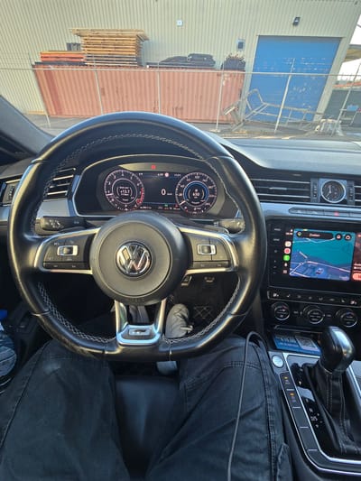 The image captures the interior of a car, specifically the driver's perspective, looking at the steering wheel, dashboard, and gear shift. The car appears to be parked outdoors, with a backdrop of industrial shipping containers and a building with a large blue door. The dashboard displays various vehicle information, including speed, engine status, and navigation. On the central infotainment screen, a map application is visible, with a route highlighted. A water bottle and a phone charger are also present in the car. The external environment suggests a location within an industrial or port area, possibly in Merelbeke, Belgium, given the context provided.