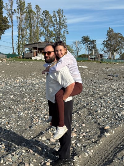 A man and a woman are on a beach in Gulripshi, Abkhazia. The woman is straddling the man's back. The man is wearing sunglasses and a light-colored shirt. The woman is wearing a white shirt. The ground is covered with stones. Trees are in the background.