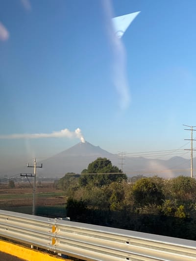 The image displays an outdoor landscape viewed through a window, evident by reflections across the upper sky and a vertical light streak on the left. In the immediate foreground, a metallic, multi-bar highway guardrail with a yellow base spans the bottom quarter of the frame. Beyond the guardrail, the midground features agricultural fields, alternating between tilled brown soil and green vegetation, extending from the left towards a line of dense green trees and shrubs that stretch across the frame. Multiple utility poles with power lines intersect the scene horizontally and vertically at various points. In the background, under a clear, bright blue sky, a large, conical stratovolcano, identified as Popocatépetl, dominates the central view. A prominent plume of white smoke or ash actively emanates from the volcano's summit, drifting horizontally to the left. Distant, hazy mountain ranges are visible to the right and behind the primary volcano. The vantage point is consistent with an area in or near Santa Ana Xalmimilulco, Mexico, looking towards the active volcano. No individuals or animals are visible.