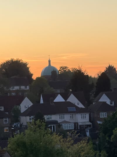 The image captures an elevated view of a residential area in High Wycombe, United Kingdom, during either sunrise or sunset. The sky dominates the left side of the frame, displaying a warm gradient of colors from a bright orange-yellow near the horizon to a paler yellow and soft blue higher up. Faint atmospheric streaks are visible across the expanse of the sky.
A distinctive dome-shaped building, possibly a church, is silhouetted against the bright sky, positioned slightly to the left of the center. It features a spire topped with a cross, and the dome itself appears to have a subtle greenish or teal tint.
The right and lower sections of the photograph are filled with a mix of residential buildings and dense tree canopy. The buildings are primarily multi-story houses with pitched roofs and various window styles, including white-framed windows and dormer windows. Many structures are partially obscured by the dark silhouettes and green foliage of numerous trees. Some windows in the buildings show faint internal illumination, suggesting habitation.
There is no visible human activity or discernible text within the image. The scene conveys a sense of tranquility and peace, characteristic of a quiet town during the golden hours of the day.