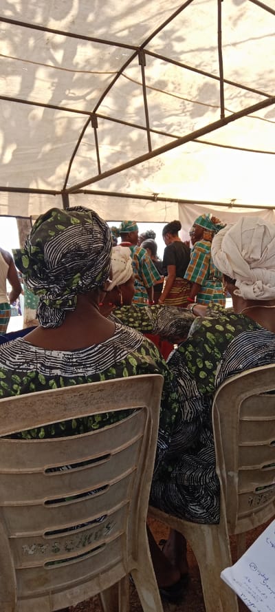 The scene depicts an outdoor gathering occurring under a white fabric canopy supported by a metal framework, in the city of Iyara, Nigeria. Sunlight casts distinct shadows of tree branches onto the canopy from above. In the immediate foreground, two individuals, likely women, are seated with their backs to the observer on off-white plastic chairs. They wear garments with a black, white, and green patterned fabric. The person on the left has a prominent headwrap (gele) in the same pattern as her attire, featuring white wavy lines and green abstract shapes. A partially visible white notebook with blue handwriting rests on the ground or a surface in the lower right.
In the mid-ground, a group of approximately eight to ten individuals is standing. Several of these individuals wear matching outfits consisting of a green, brown, and white patterned fabric, possibly plaid or striped, along with matching headwraps. One person in this group, centrally positioned, wears a dark short-sleeved top and a horizontally striped skirt with hues of brown, cream, and possibly red, and has dark hair pulled back. The individuals in the mid-ground appear to be facing away or in profile, engaging with each other or observing an unseen focal point. The ground beneath the chairs is a reddish-brown color, consistent with unpaved earth.