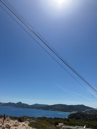The image depicts a scenic view in Cala Rajada, Spain. The foreground features a rocky terrain with sparse vegetation. A figure is visible at the bottom right corner. In the middle ground is a bay with calm, dark blue water. Across the bay, there's a mountain ridge covered in trees, dotted with buildings. The sky is clear blue with some power lines stretching across and two streaks suggesting aircraft trails. The sun is visible in the top left corner.