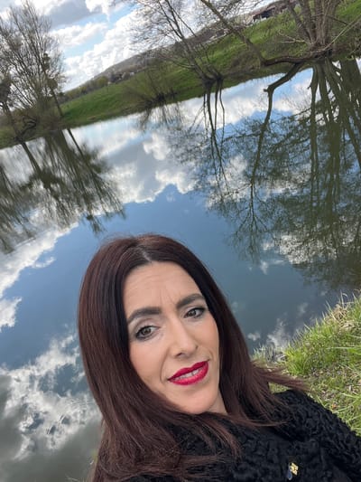The image shows a woman with long, dark hair, wearing a black textured coat, posing for a selfie. She has fair skin, red lipstick, and appears to be smiling slightly. The background features a body of water reflecting the sky, clouds, and trees, suggesting the picture was taken near a river or lake. Based on the location provided, the scene is likely in Gavisse, France, where there are numerous waterways and natural landscapes. The environmental elements suggest an outdoor setting with calm waters mirroring the sky and surrounding vegetation.