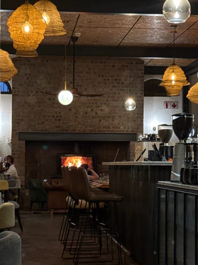 Indoor shot of a restaurant, potentially in Paarl, South Africa. The ceiling features unique lighting fixtures. A fireplace is visible in the scene. Diners are present, with one man in a white shirt clearly visible. Furniture, including chairs and stacked bar stools, is part of the setting.