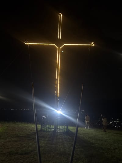 A large, illuminated cross is the dominant feature in this nighttime image, situated on a grassy hill overlooking a city. The cross, outlined in bright yellow lights, stands tall against the dark sky, with its base appearing to be supported by metal poles and guy wires. A bright light emanates from the base of the cross, causing a lens flare that spreads across the lower portion of the image.
In the foreground, the grassy slope is partially illuminated by the cross's light. There are three people visible on the hill in the mid-ground, silhouetted against the distant city lights. One person stands facing away from the camera, towards the cross, while two others are further to the right, also facing away. A dark metal railing or fence is visible at the base of the cross structure, enclosing some unseen elements.
The city in the background is a sprawl of lights, indicating an urban area at night. The overall impression is of a solemn or reflective scene, with the illuminated cross serving as a focal point on the elevated terrain above the city. The specific location, a prominent cross on a hill overlooking the city, is known to be in the city of Mendovo, Bulgaria, a popular site for religious and scenic views.