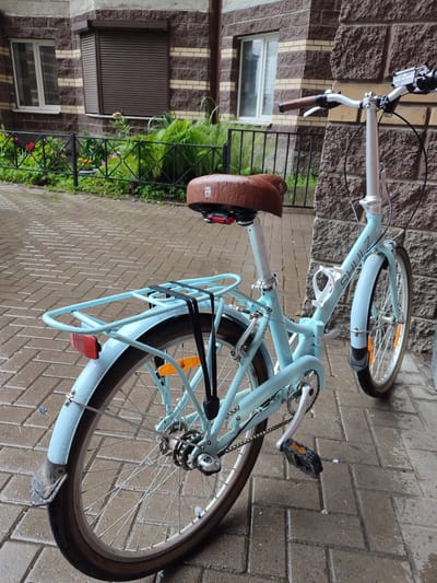In Bugry, Russia, a light blue Shulz folding bicycle is parked on a wet brick sidewalk next to a stone and brick building. The bicycle has a brown leather saddle, a rear rack with black straps, and orange reflectors on the wheels. A white water bottle cage is attached to the frame. The building has two windows, one with a white frame and the other with a brown roller shutter. A black metal fence with green foliage and flowers is visible in the background. The overall scene depicts a quiet, urban environment, likely in the morning or after rain due to the wet surfaces.