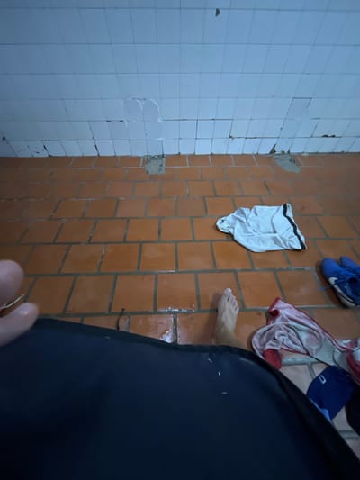 The image appears to be taken in a bathroom setting. The walls are tiled, with white tiles on one side and brown tiles on the other. There's damage to the tiled walls in several places. The floor is also tiled with brown tiles. On the brown tiled wall, there are objects hanging: a pair of blue running shoes, a pair of white athletic shorts, and a red and white striped towel along with a blue athletic hat. A person is present, standing on the floor. The person's foot is visible. A large black garment is positioned close to the camera, obscuring some of the scene. The photograph's perspective is at a low angle, looking upwards. There's no definitive information within the image that confirms its location within the city of Moreno, Argentina.